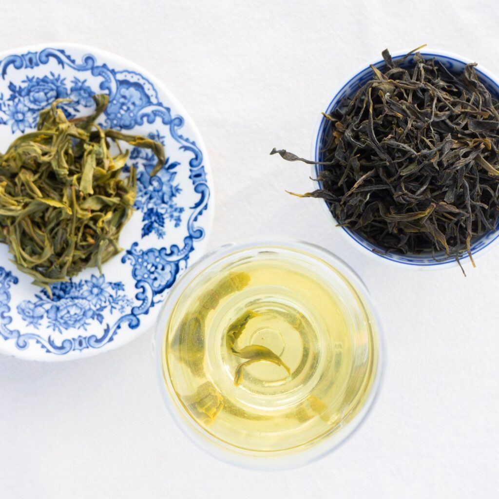 A glass of brewed tea alongside green tea leaves in decorative bowls.