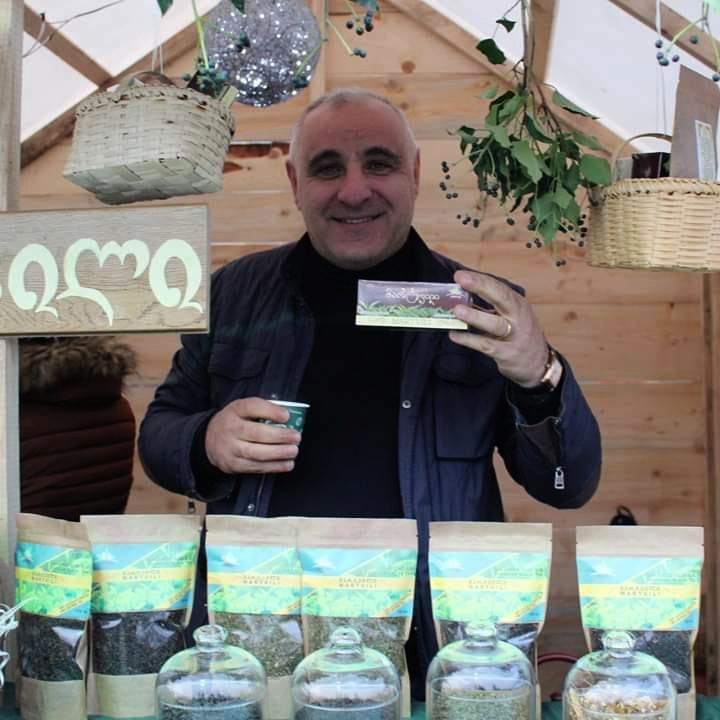 Gela smiling and holding a box of Martvili Tea in front of a display of tea bags at a market stall.