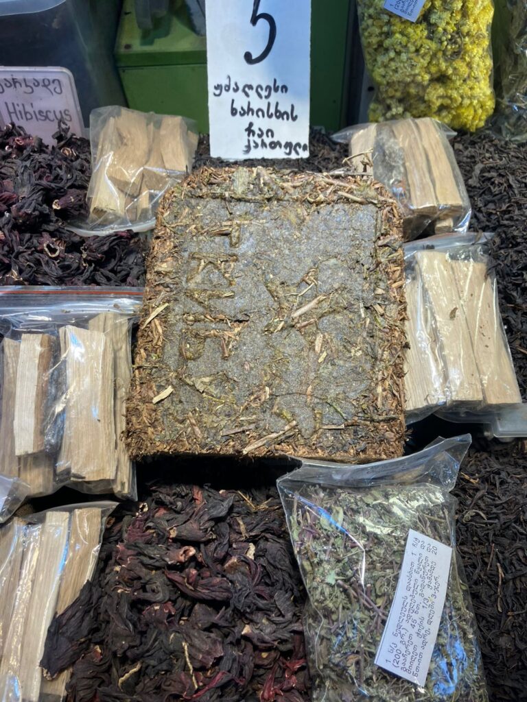 Brown square tea brick on display among dried herbs and loose leaves at a market stall, with plastic bags containing herbs and writing on signs